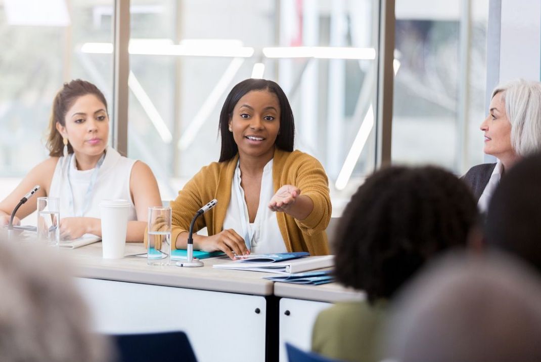 Three people sitting at a table, the person in the middle speaking with their hand out in gesture.