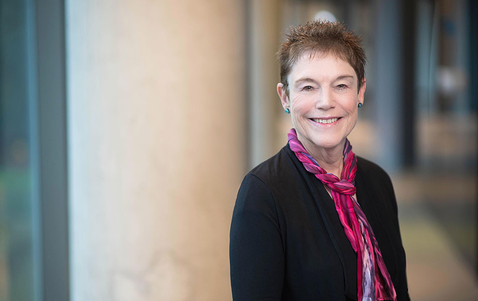 A photo of Bonny Norton smiling, standing beside a concrete pillar in a hallway. She is wearing a black jumper and a pink scarf