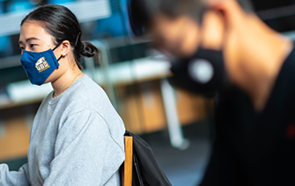 An image of students wearing masks, working at computers. One is wearing a blue mask with the UBC logo and a grey sweater. The other is wearing a black sweater.