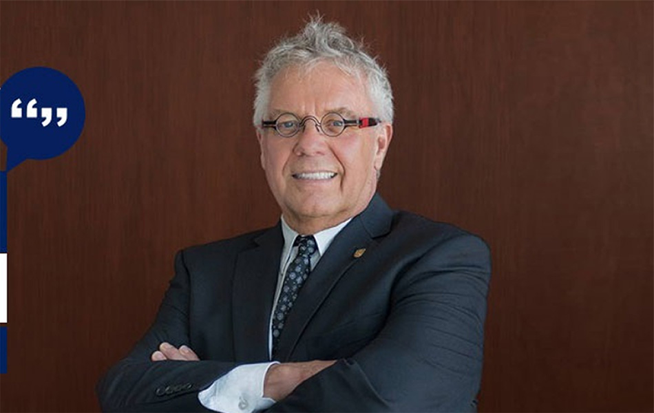 An image of Dean Blye Frank smiling, standing with arms crossed against a wooden panel wearing a suit and glasses