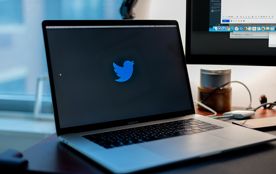 An image of laptop computer open with the Twitter icon displayed, and a projector screen in the background