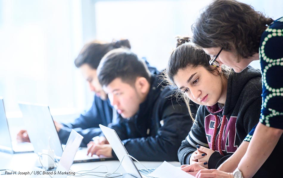 A teacher leans over her students work, both staring down at the paper, while students work on their laptops in the background