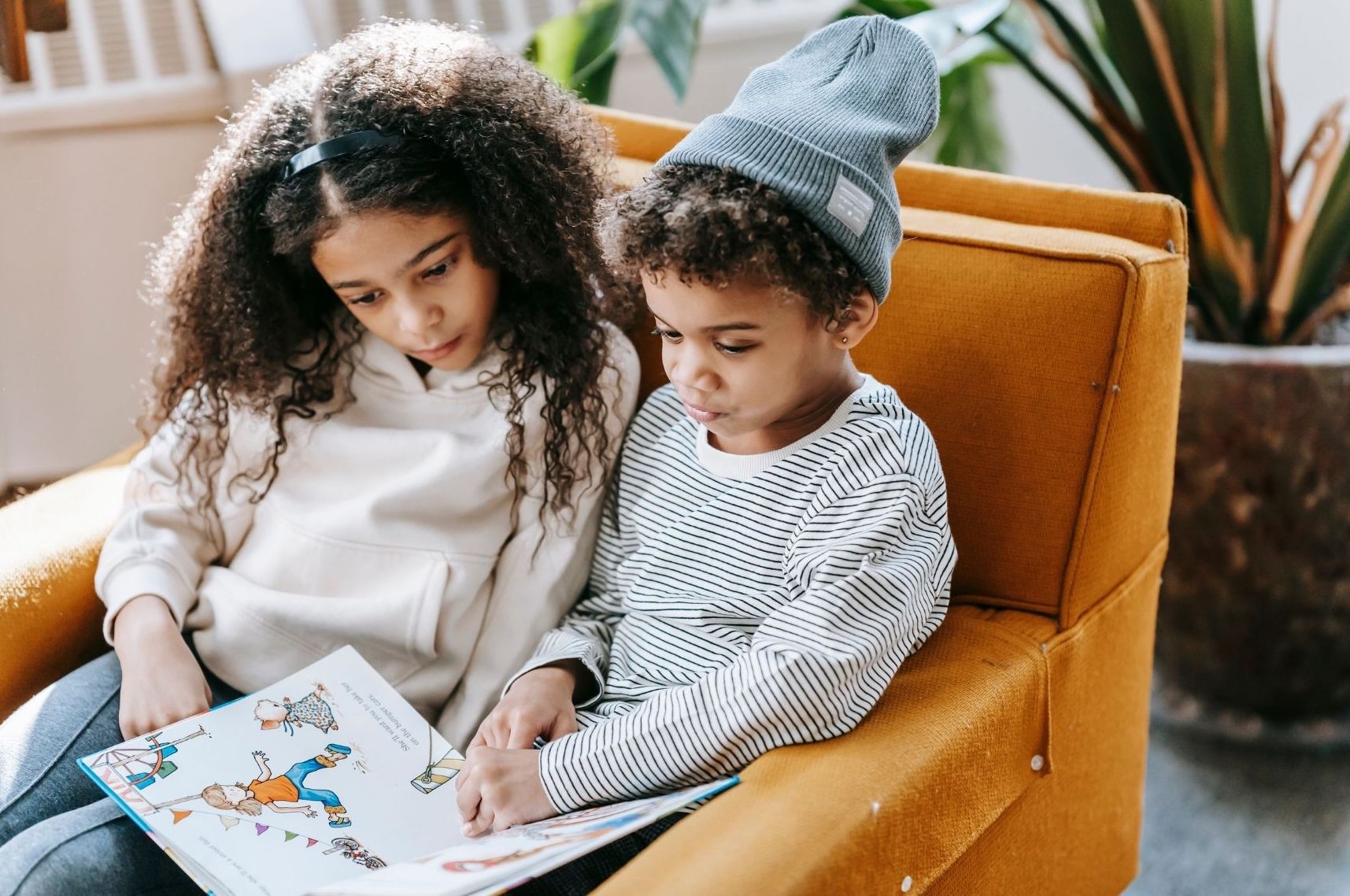 Two young children sitting together in an armchair, reading a book with pictures and text.