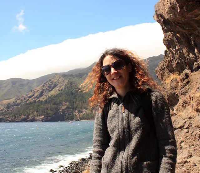 A photo of Dr. Dianne Newbury standing on the shores of Robinson Crusoe Island, with the ocean and part of the island in the background. She is wearing a grey jumper and sunglasses.