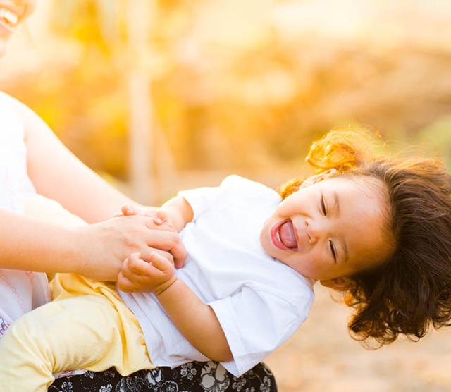 An image of a parent holding a laughing child, who is leaning almost horizontal, with sunlight in the background