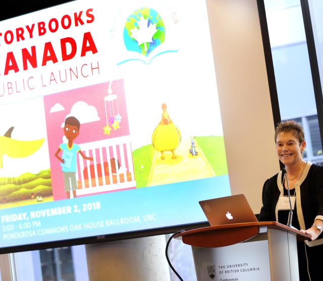 An image of Bonny Norton standing at a podium in front of a projector screen, at the launch of Storybooks Canada, 