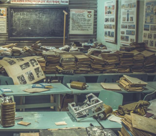 A picture of a cluttered classroom with a blackboard at the front, green tables and desks piled with books and papers, and posters on the walls.