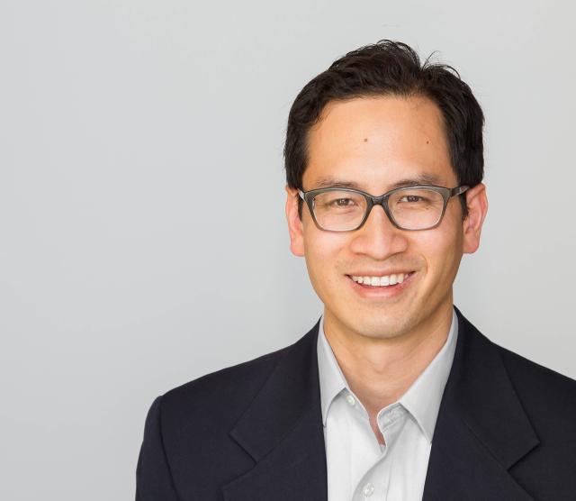 A headshot of Professor Edward Chang, smiling, wearing glasses, and standing against a grey background.