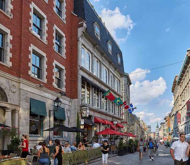 An image of a downtown Montreal street lined with flags, with a brick building on the left, people sitting on the street at tables, and people walking down the street