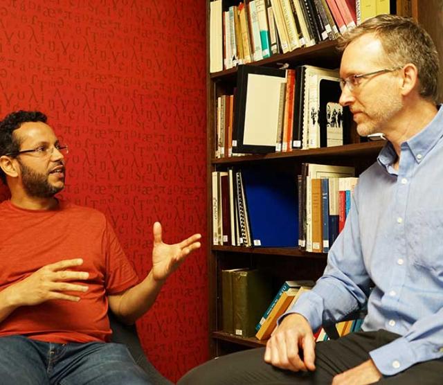 A picture of assistant professor Muhammad Abdul-Mageed speaking and Professor Bryan Gick listening against a red wall, with a bookcase in the background