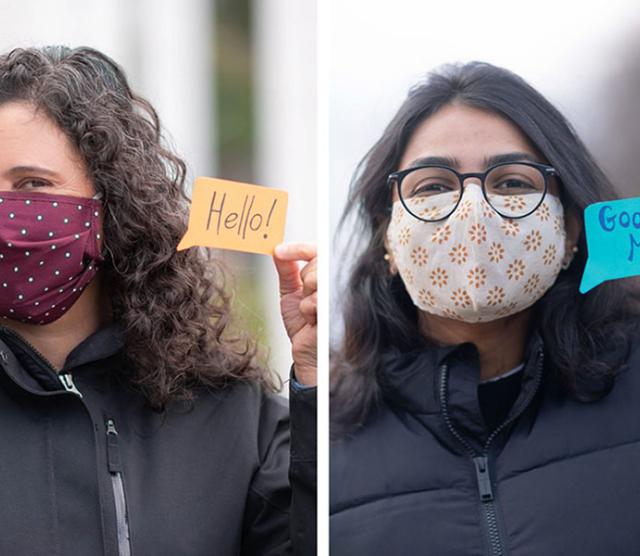 An image of two students wearing facemasks, and jackets, standing outside, holding up paper in the shape of speech bubbles with greetings written on the paper