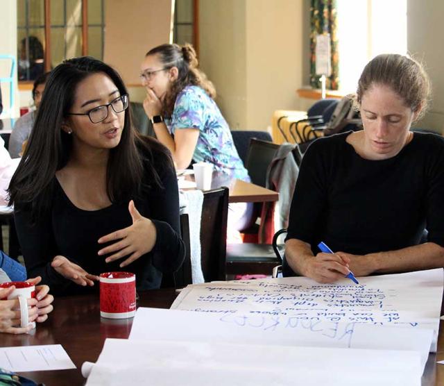 Two language sciences members sit at a table, discussing research at a recent member event.  One of them, a woman with long dark hair, glasses, and a dark-colored long-sleeved shirt, gestures while speaking, while the other, a woman with light-coloured hair pulled back in a ponytail, wearing a dark-coloured short-sleeved shirt,  writes down notes on a large flipchart on the table in front of them.  Other language sciences members can be seen conversing in the background.