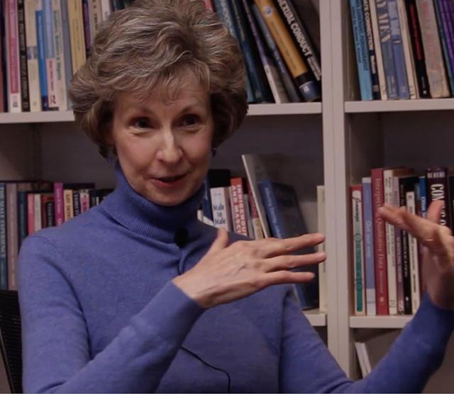 A picture of Professor Janet Jamieson wearing a purple sweater sitting in front of a bookcase, midway through the ASL sign for Star Wars