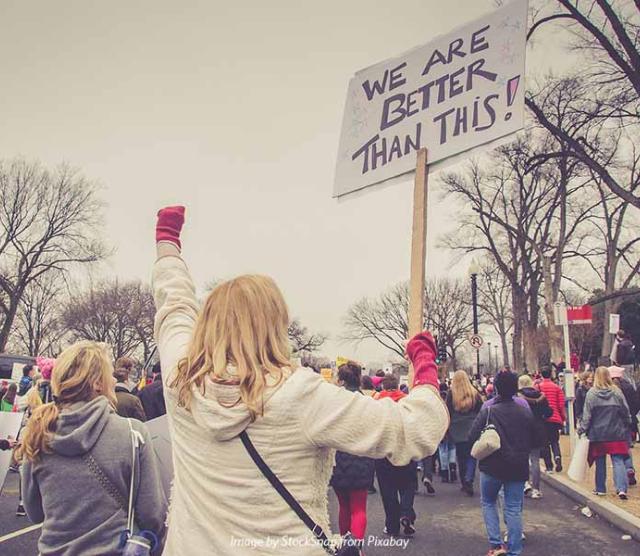 A photo from the back of protestors marching along a road, with a blonde woman in the foreground, her fist in a red glove and raised, holding a sign saying We are better than this
