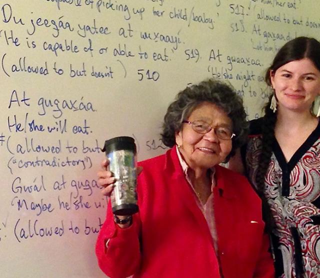 A picture of Elder Keiyishí Bessie Cooley, wearing a red jumper and raising a coffee travel mug, and Heather Burge wearing a dress with her arm around Elder Cooley. Both are standing in front of a whiteboard with Lingít and English on it
