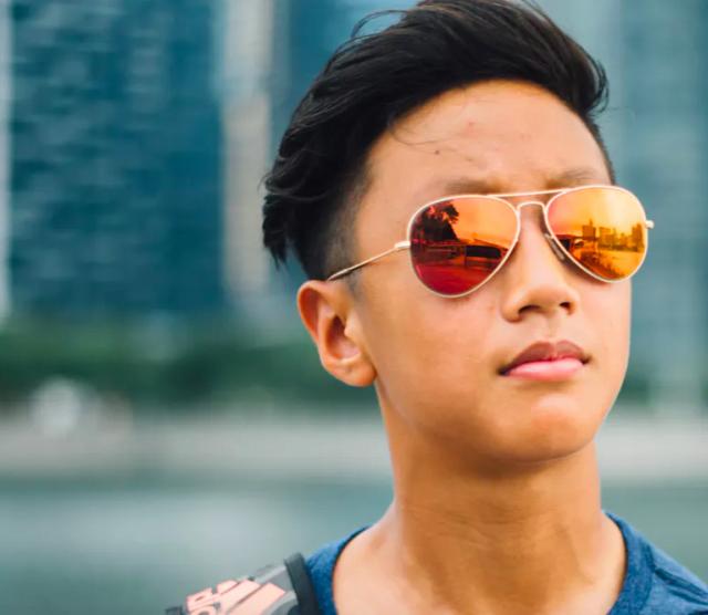 A picture of a young person of Asian heritage wearing sunglasses and standing in front of a harbour