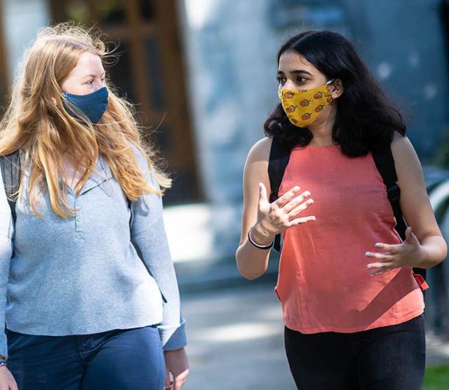 An image of two students walking on UBC campus, wearing masks and with long hair. One student is gesturing as they talk.
