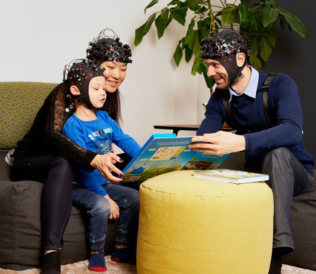 An image of parents reading a book to a child. Everyone is wearing EEG caps.