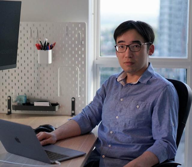 Keyi Tang sitting at a desk in front of a latop and a window with a cityscape. He is wearing a blue shirt and glasses