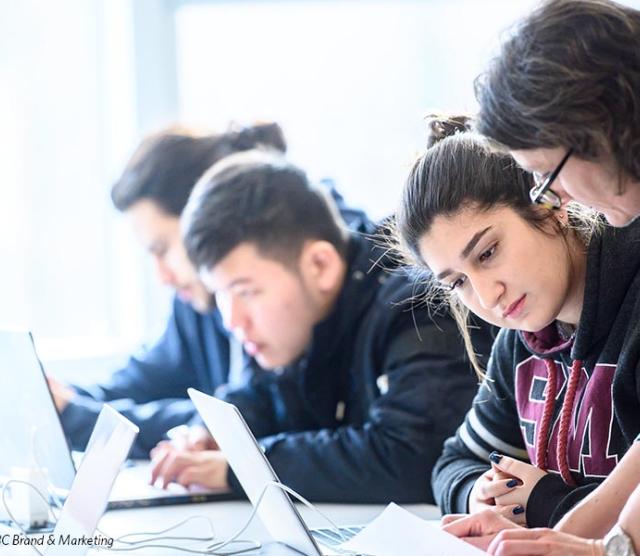 A teacher leans over her students work, both staring down at the paper, while students work on their laptops in the background
