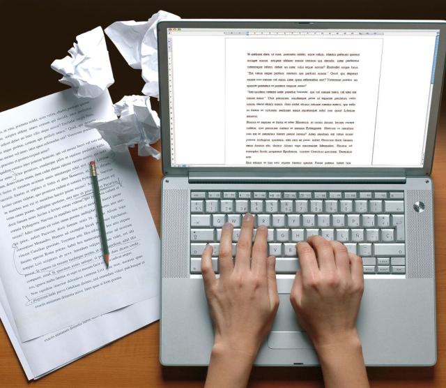 Desk with an open laptop and hands typing. Loose papers scattered around.