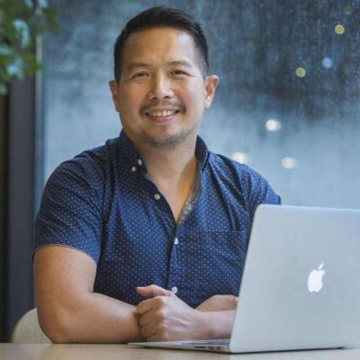 A picture of Ron Darvin smiling, sitting in front of a laptop with arms crossed on the table.