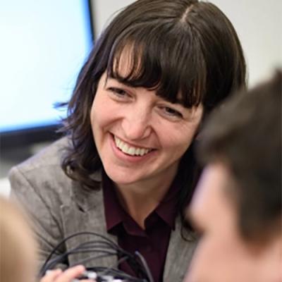 An image of Lauren Emberson smiling as she listens to someone, sitting in front of a projector screen. Part of someones head is blurred in the right foreground