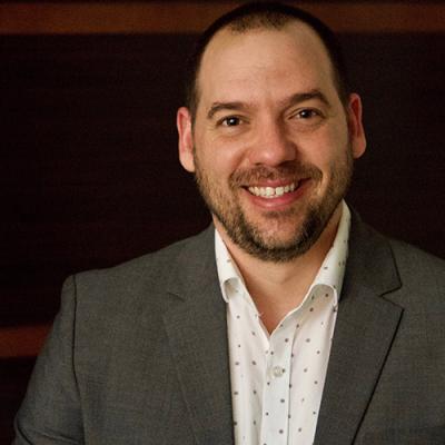 A headshot of Dr Labrada dressed in a grey jacket and white shirt, standing against a dark background and smiling