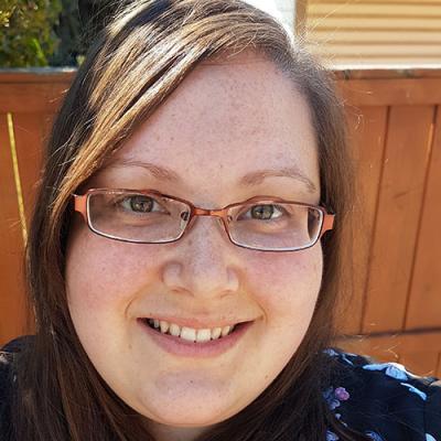 A headshot of Brianna Peacey smiling, wearing glasses, with a brown fence in the background