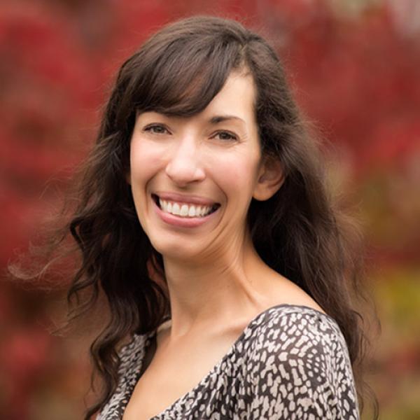 A headshot of Dr. Kathryn Accurso standing side on, smiling, in front of a tree with red leaves