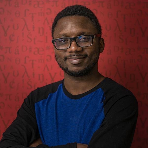 A headshot of Samuel Akinbo with arms folded, wearing glasses, standing against a red background