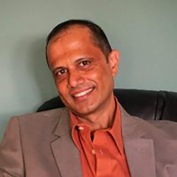 A headshot of Reginald D'Silva smiling, and wearing an orange shirt under a tan jacket, sitting in a chair against a green wall