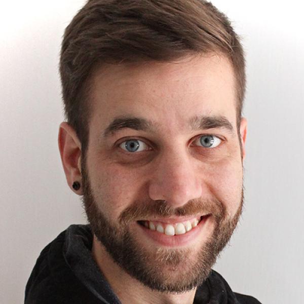 A headshot of Félix Desmeules-Trudel wearing a black hoodie and a black ear plug, smiling at the camera against a white background