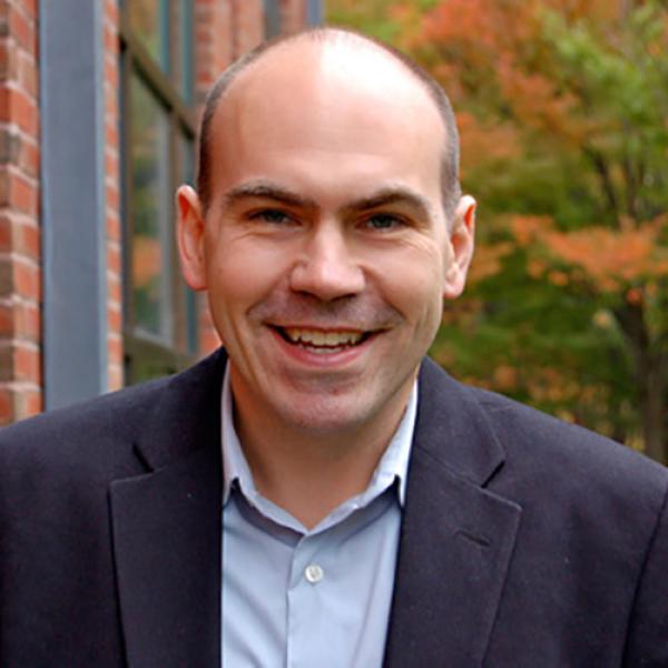 A headshot of Jonathan Peelle smiling, wearing a dark blue jacket and a light blue shirt, standing oustide a brick and glass building with fall trees in the background