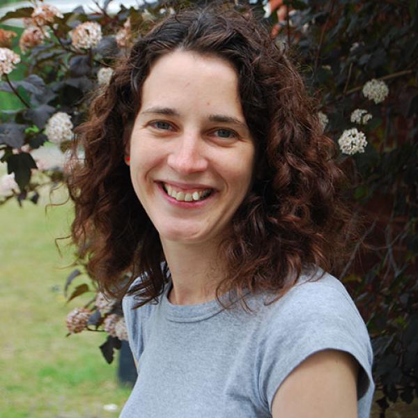 A headshot of Dianne Newbury smiling at the camera, standing outside in front of a flowering bush, and wearing a light blue t-shirt