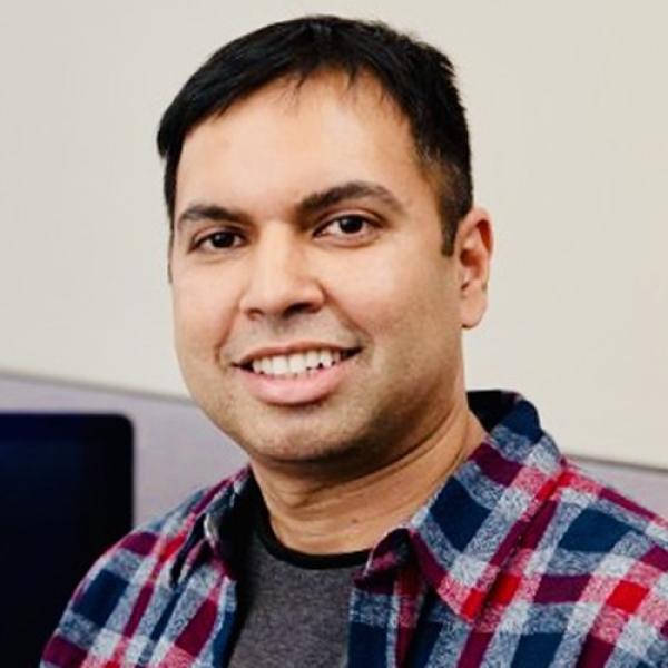 A headshot of Dr. Saif Mohammad smiling into camera, wearing a plaid shirt over a grey shirt, standing in front of a beige background